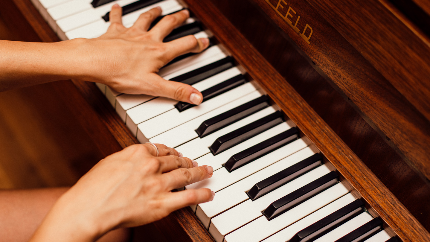 Two hands over an acoustic piano keyboard, left hand playing Eb major chord