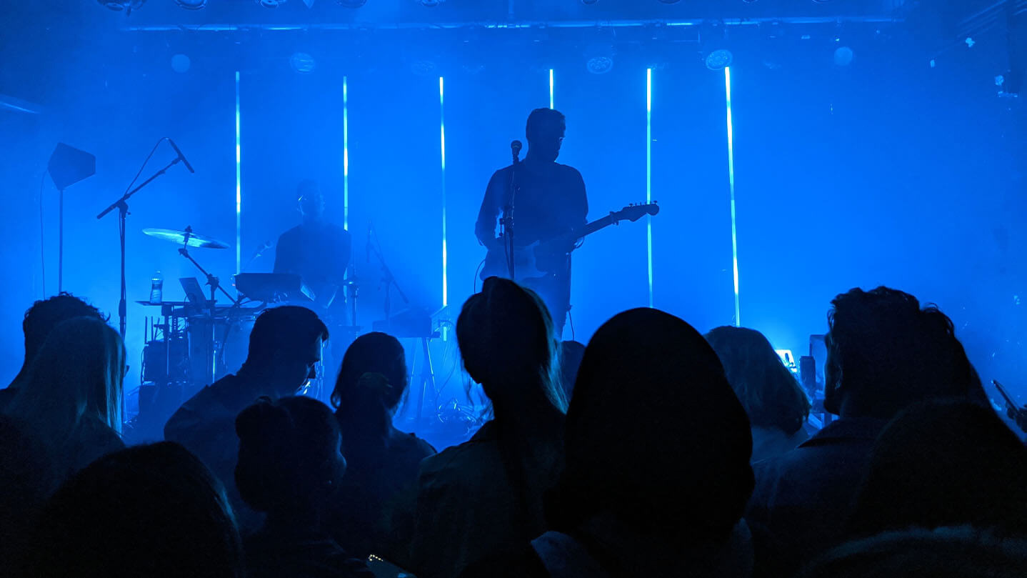 Photo taken amidst a crowd of people, watching a guitarist and drummer on stage. Blue stage lighting.