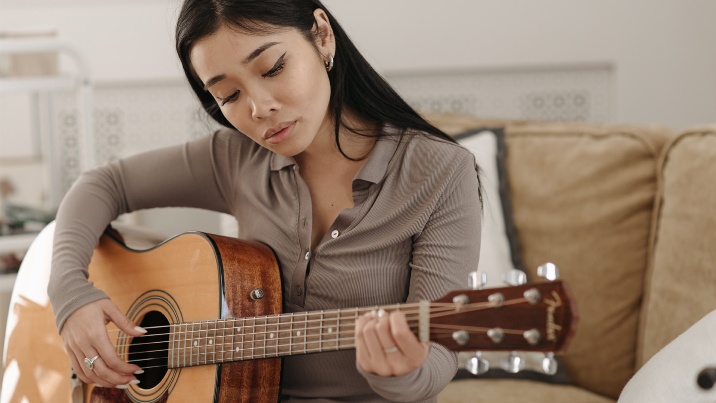 Woman seated on couch, playing a Fender acoustic guitar. 
