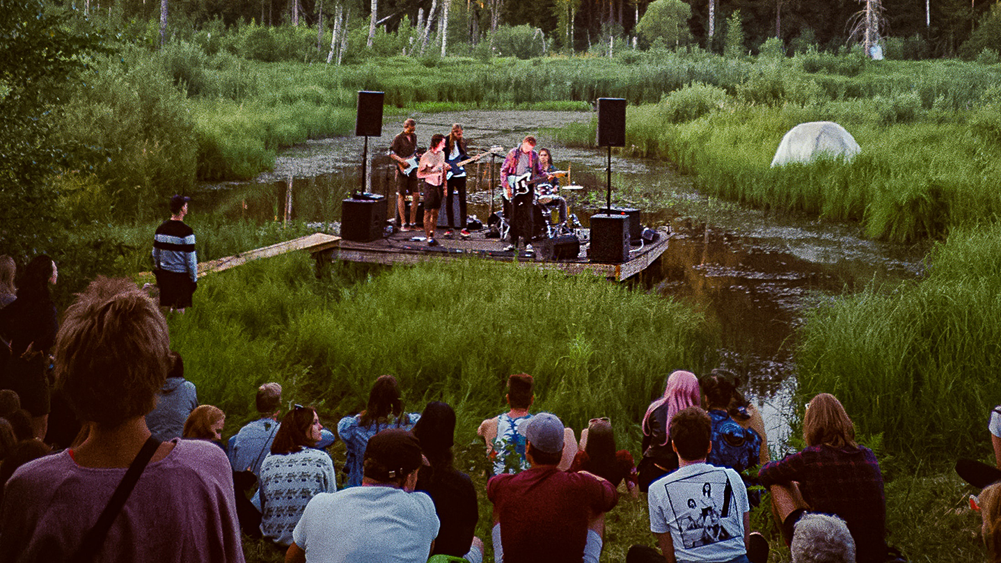 6-piece band playing on an outdoor stage built into a swamp - small crowd sits on the shore, watching the show.