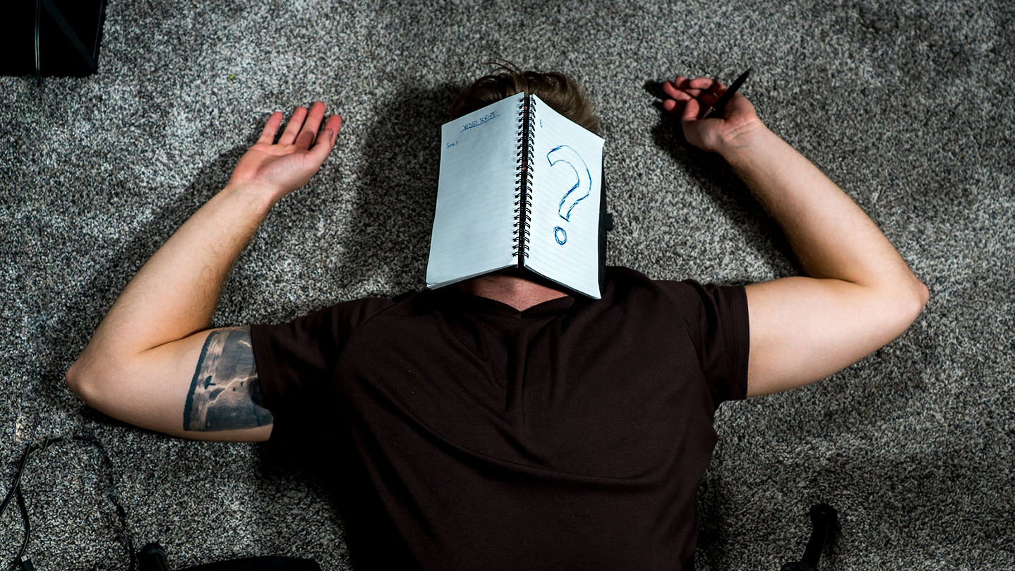Person lying on a carpet with their hands over their head and an open notebook laying on their face