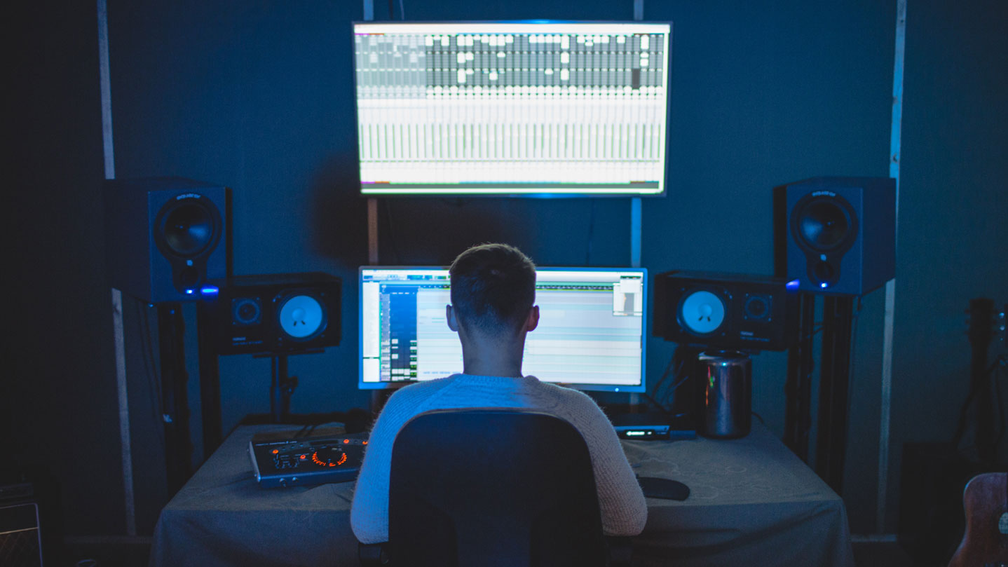 Person sits at a desk in front of a recording setup with studio monitors and a computer