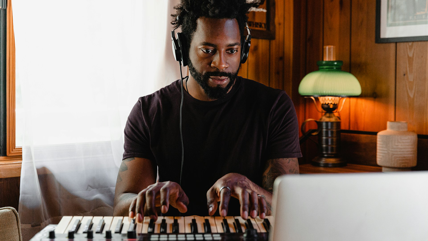 Bandzoogle blog: 3 ideas to help musicians stop the grind and thrive more. Image of a man sitting at a piano keyboard, playing with headphones on, and looking into a laptop screen.