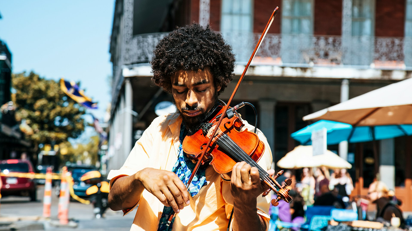 Bandzoogle blog: 6 tips on songwriting with new instruments. Image of a man playing violin on a busy, sunny, city street corner.